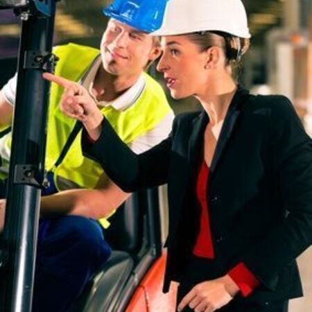Eine Frau in einem Bauhelm und ein Mann in einem Anzug stehen neben einem Gabelstapler und Unterhalten sich